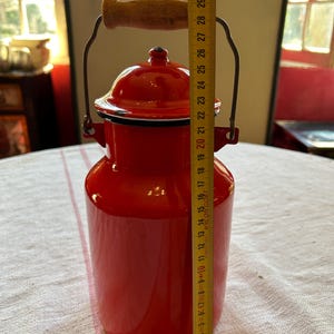 Könnte beinhalten: Rote Emaille-Milchkanne mit weißem Rand und Holzgriff. Die Kanne ist 71 cm hoch.