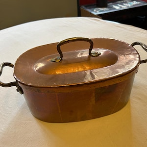 May include: Oval copper cooking pot with a lid and two side handles. The pot has a central handle on the lid. The copper has a warm, reddish-brown colour, and the handles appear to be made of a darker metal.