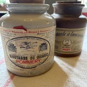 May include: Two ceramic jars of mustard. The jar on the left is white with a brown lid and a label that reads "Moutarde de Meaux Pommery" in black and red lettering. The jar on the right is brown with a brown lid and a label that reads "Moutarde à l'ancienne" in black lettering.