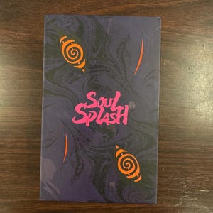Puede incluir: Una caja morada con un patr&oacute;n de m&aacute;rmol y el texto "Soul Splash" en letras rosas. La caja tiene dos dise&ntilde;os en espiral naranja y negro en la parte delantera.