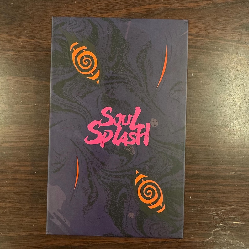 Puede incluir: Una caja morada con un patr&oacute;n de m&aacute;rmol y el texto "Soul Splash" en letras rosas. La caja tiene dos dise&ntilde;os en espiral naranja y negro en la parte delantera.