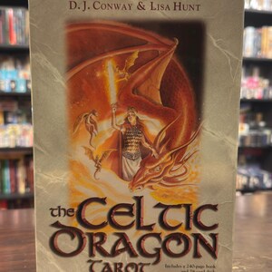Puede incluir: La caja del mazo de tarot Celtic Dragon presenta un dragón rojo con una mujer sosteniendo una espada. La caja es beige con texto marrón que dice "The Celtic Dragon Tarot" y "Incluye un libro de 240 páginas y una baraja de 78 cartas."
