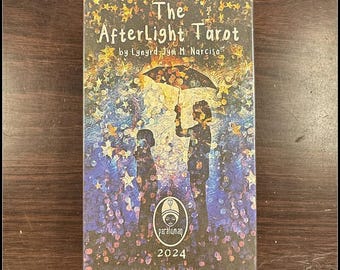 May include: A tarot card box titled "The Afterlight Tarot" by Lynyrd-Jym M. Narciso. The box shows an illustration of two figures under an umbrella against a starry, colourful backdrop. The year 2024 is printed at the bottom of the box.