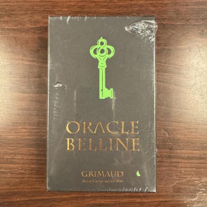 Puede incluir: Una caja negra sellada con las palabras "ORACLE BELLINE" en letras doradas y un gráfico de llave verde. La marca "GRIMAUD" está impresa debajo del título. La caja está envuelta en plástico transparente.