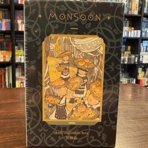 Puede incluir: Una caja negra con un borde dorado y la palabra "MONSOON" en letras doradas. La caja presenta una ilustración de personas que llevan cestas de pan. El texto "Anti-collision box" y "防碰撞盒" está impreso en la parte inferior de la caja.