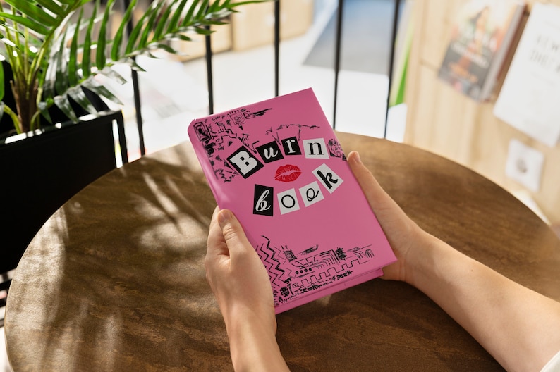 Puede incluir: Una libreta rosa con las palabras "Burn Book" escritas en letras negras con una marca de l&aacute;piz labial rojo. La libreta est&aacute; decorada con garabatos en blanco y negro.