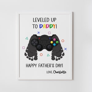Pode incluir: Uma impressão emoldurada com o texto "LEVELED UP TO DADDY! HAPPY FATHER'S DAY! LOVE. Charlotte." Um controle de videogame preto está no centro, com pegadas em ambos os lados. Formas coloridas circundam o controle.
