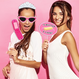 May include: Two women pose for a photo with pink and white photo booth props. One woman holds a pink flip phone prop and wears a tiara and pink sunglasses. The other woman holds a sign that says "Is butter a carb?" with a stick of butter illustration.