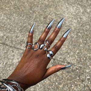 Może przedstawiać: Dłoń ozdobiona wieloma srebrnymi pierścionkami o różnych wzorach, w tym otwartymi okręgami i gładkimi, zaokrąglonymi kształtami. Palce pomalowane są długimi, metalicznymi srebrnymi paznokciami. Na nadgarstku widoczna jest srebrna bransoletka.