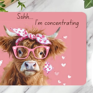 May include: A pink mouse pad with a cartoon Highland cow wearing pink glasses and a pink bandana with white hearts. The cow is looking at the camera and the text "Sshh... I'm concentrating" is written above the cow.