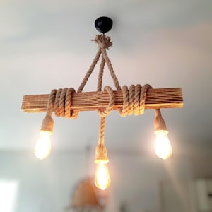 Könnte beinhalten: Ein rustikaler Holz-Kronleuchter mit drei Edison-Glühbirnen. Der Holzbalken ist mit dickem Seil umwickelt, und die Leuchte hängt an einer Deckenhalterung. Die Glühbirnen sind beleuchtet und erzeugen einen warmen Schein.