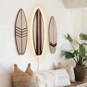 Peut inclure: Trois décorations murales en forme de planche de surf. Les pièces de gauche et de droite présentent des motifs géométriques marron foncé sur une finition en bois clair. La pièce centrale est une planche en bois foncé avec un contour LED lumineux.