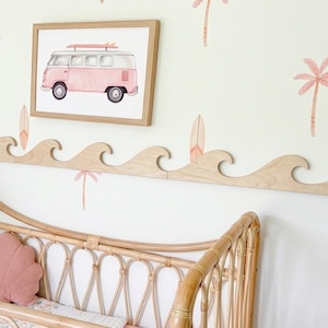 Könnte beinhalten: Ein Kinderbett aus Rattan mit einem rosa muschelförmigen Kissen und einer gemusterten Decke. Über dem Bett befindet sich eine hölzerne Wandleiste in Wellenform unter einem gerahmten Bild eines rosa Vintage-Vans mit einem Surfbrett auf dem Dach.