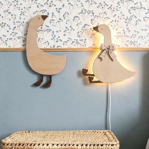 Op de afbeelding: Twee houten wanddecoraties in de vorm van een gans. De linker gans staat, terwijl de rechter gans zit en is voorzien van een geïntegreerde warme LED-lichtbron achter het lichaam, een klein stoffen strikje om de nek en een wit snoer.