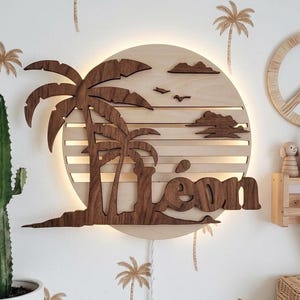 May include: Wooden wall art featuring a tropical scene with palm trees, clouds, and the name "Léon". The art is illuminated with a warm glow, creating a sunset effect. Palm tree decals decorate the white wall.