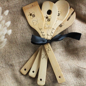 Può includere: Un set di sei utensili da cucina in legno, legati con un nastro nero. Ogni utensile presenta un'ape e la parola "Bee" o "happy" incisa sul legno chiaro. Include una spatola, un cucchiaio e una forchetta.