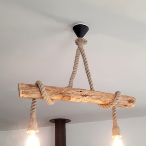 Pode incluir: Uma luminária rústica de madeira suspensa do teto por uma corda grossa bege. Duas lâmpadas estilo Edison pendem abaixo da viga. A luminária é fixada a uma montagem de teto preta e branca, criando uma estética natural e náutica.