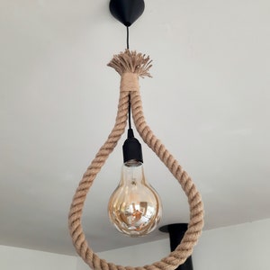 Peut inclure: Un luminaire suspendu en corde naturelle avec une seule ampoule. La corde est tissée en forme de larme et pend d'un support de plafond noir.