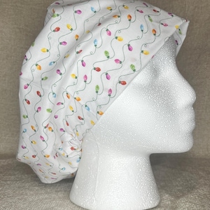 Puede incluir: Un gorro quirúrgico blanco con un patrón repetido de luces navideñas de colores. Las luces son en tonos de rojo, amarillo, verde, naranja y azul, conectadas por un cable verde. El gorro se muestra en una cabeza de maniquí blanca.