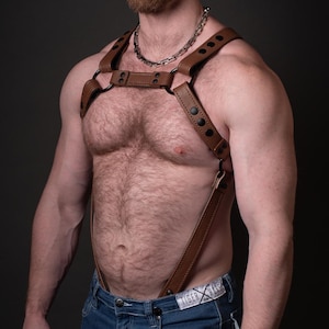 May include: A man wearing a brown leather harness with black buckles and a silver chain necklace. He is wearing blue jeans with a white label that says "Tiger X Rise".