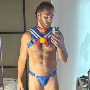 May include: Blue and white cosplay outfit with a red collar and a large bow. The outfit includes a harness and briefs. The person is holding a phone and looking in a mirror.