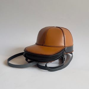 May include: A brown leather baseball cap purse with a black zipper and strap.