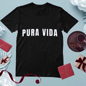 May include: Black t-shirt with the white text "PURA VIDA". The shirt is surrounded by decorative elements, including wrapped gifts and a cup of tea on a dark brown plate. The overall theme suggests a lifestyle or gift item.