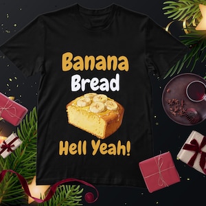 May include: Black t-shirt with the words "Banana Bread Hell Yeah!" in yellow and white. The shirt features an illustration of a slice of banana bread topped with banana slices. The image is surrounded by holiday decorations.