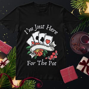 May include: A black short-sleeved t-shirt featuring a graphic of four playing cards, a red flower, and poker chips. The text "I'm Just Here For The Pot" is printed in white above and below the central illustration in a bold, clean font.