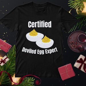 Può includere: T-shirt nero con la scritta "Certified Deviled Egg Expert" e l'illustrazione di due uova ripiene con ripieno giallo. Decorazioni festive sullo sfondo.