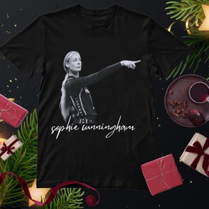 Puede incluir: Una camiseta negra de manga corta con un estampado fotográfico monocromático de una jugadora de baloncesto señalando hacia un lado, con el nombre Sophie Cunningham escrito en una fuente cursiva blanca debajo de la imagen en el frente.