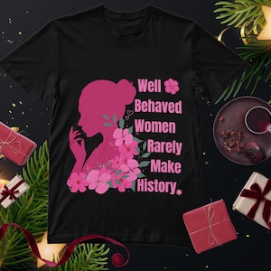May include: Black t-shirt featuring a pink silhouette of a woman and pink flowers. The text on the shirt says "Well Behaved Women Rarely Make History". The image is decorated with festive elements.