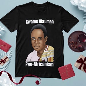 May include: Black t-shirt featuring a portrait of Kwame Nkrumah with the text "Kwame Nkrumah" above the image and "Pan-Africanism" below. The portrait is a stylized illustration. The shirt is surrounded by decorative elements.