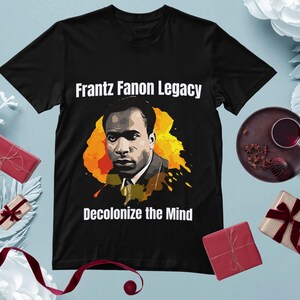 May include: Black t-shirt featuring a portrait of Frantz Fanon with the text "Frantz Fanon Legacy" and "Decolonize the Mind" in white. The portrait is set against a yellow and orange splash design. A thought-provoking graphic tee.