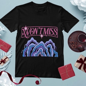 Può includere: T-shirt nero con la scritta "WON'T MISS" in rosa e una grafica di montagna nei toni del blu e del viola. La maglietta è presentata con regali e una bevanda su una superficie azzurra.
