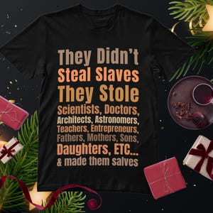 May include: Black t-shirt with the text "They Didn't Steal Slaves They Stole Scientists, Doctors, Architects, Astronomers, Teachers, Entrepreneurs, Fathers, Mothers, Sons, Daughters, ETC... & made them salves". The shirt is surrounded by holiday decorations.