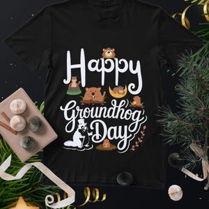 May include: Black t-shirt featuring the text "Happy Groundhog Day" in white, with illustrations of groundhogs. The design includes various groundhog illustrations. The shirt is made of a soft material.