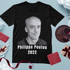 Puede incluir: Camiseta negra con un retrato en escala de grises de Philippe Poutou y el texto blanco "Philippe Poutou 2022". La imagen está rodeada de elementos decorativos, incluyendo regalos y una bebida.