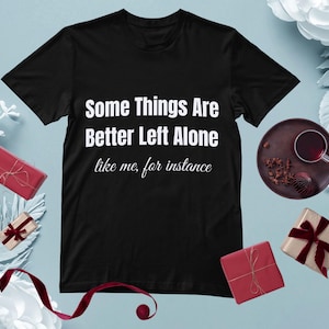 Puede incluir: Camiseta negra con el texto blanco "Some Things Are Better Left Alone like me, for instance". La camiseta está rodeada de pequeños regalos envueltos con cintas rojas y una taza de té en un plato marrón oscuro.