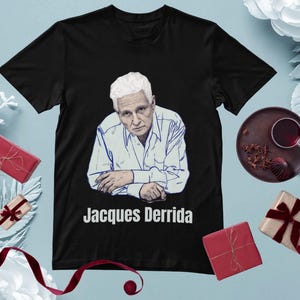 Peut inclure: T-shirt noir avec un portrait de Jacques Derrida en chemise blanche et le texte "Jacques Derrida" en dessous. L'image est entourée d'éléments décoratifs, notamment des cadeaux emballés et une tasse de thé.