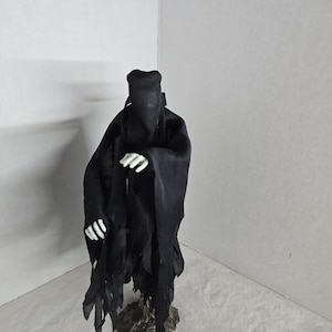 May include: A black figure with a hooded cloak and tattered edges, standing on a textured base. The figure has white skeletal hands. The overall design suggests a gothic or fantasy theme.
