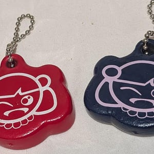 May include: Two keychains with a cartoon girl design. One keychain is red with a white outline, and the other is navy blue with a pink outline. Each keychain has a silver chain.