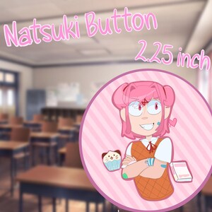 May include: A circular pin-back button measuring 2.25 inches in diameter. The design features a pink-haired character with a white shirt, orange vest, and red ribbon, holding a cat-themed cupcake. The text Natsuki Button is printed above.