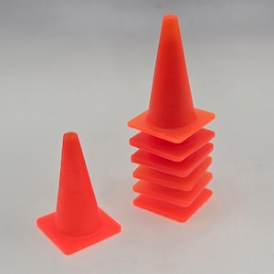May include: A stack of eight orange traffic cones, with one smaller cone in front of the stack. The cones are made of a bright orange plastic.