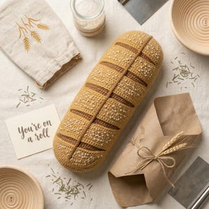 Puede incluir: Un cojín con forma de pan tejido a ganchillo en tonos beige y marrón, con una superficie texturizada y rayas decorativas. La imagen incluye una toalla de lino con bordado de trigo, una tarjeta pequeña con el texto "You're on a roll".