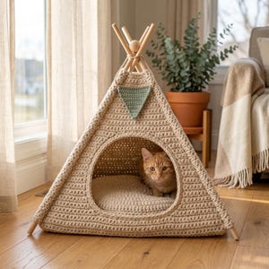 May include: A beige crochet cat teepee with wooden poles and a green triangle detail. An orange tabby cat is inside on a beige cushion. The teepee is on a wooden floor.