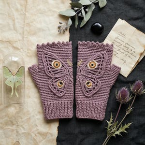 May include: A pair of mauve fingerless gloves with a crocheted butterfly design. The butterfly has detailed eye-like patterns in shades of gold and brown. The gloves are displayed on a textured surface with a handwritten note.