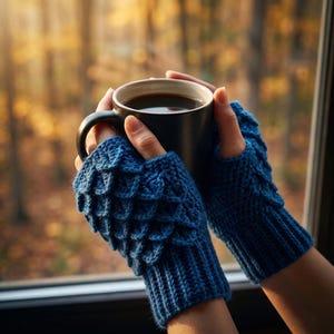 May include: A person holding a black mug of coffee, wearing blue fingerless gloves. The gloves have a textured, scale-like design on the back of the hand and a ribbed knit cuff. The background is a blurred view of a forest.