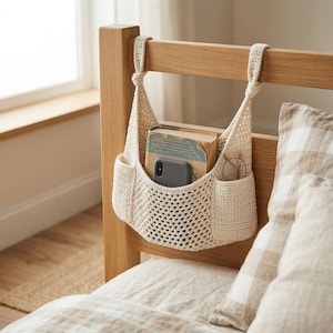 May include: A cream-colored, crocheted bed organizer hanging from a wooden bed frame. The organizer has a large central pocket holding a book and a phone, with smaller side pockets for glasses. The bed is covered with a white sheet and a patterned pillow.