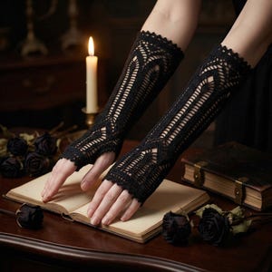 Pode incluir: Luvas de braço de crochê preto de tecido aberto com uma borda ondulada. As luvas de braço apresentam um design geométrico e são exibidas em uma mesa de madeira com um livro, rosas pretas e uma vela acesa.